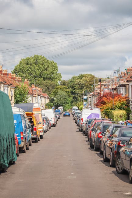 A quiet residential street with a mix of terraced houses and lush green trees lining the pavements under a cloudy sky. Parked vehicles, including various vans, small trucks, and cars, occupy both sides of the narrow, tarmac road. Some of the vans are covered with protective fabric or plastic wrap, indicating ongoing packing or loading activities typical of home relocation or furniture transport. In the foreground, a white van belonging to Man and Van Highbury is visible, positioned near the curb, likely in the process of loading or unloading furniture and packing materials such as cardboard boxes, plastic wraps, and blankets. The image captures a scene representative of the logistical steps involved in house removals or packing and moving services within an urban environment, highlighting the careful organization and vehicle arrangement central to efficient relocation efforts.