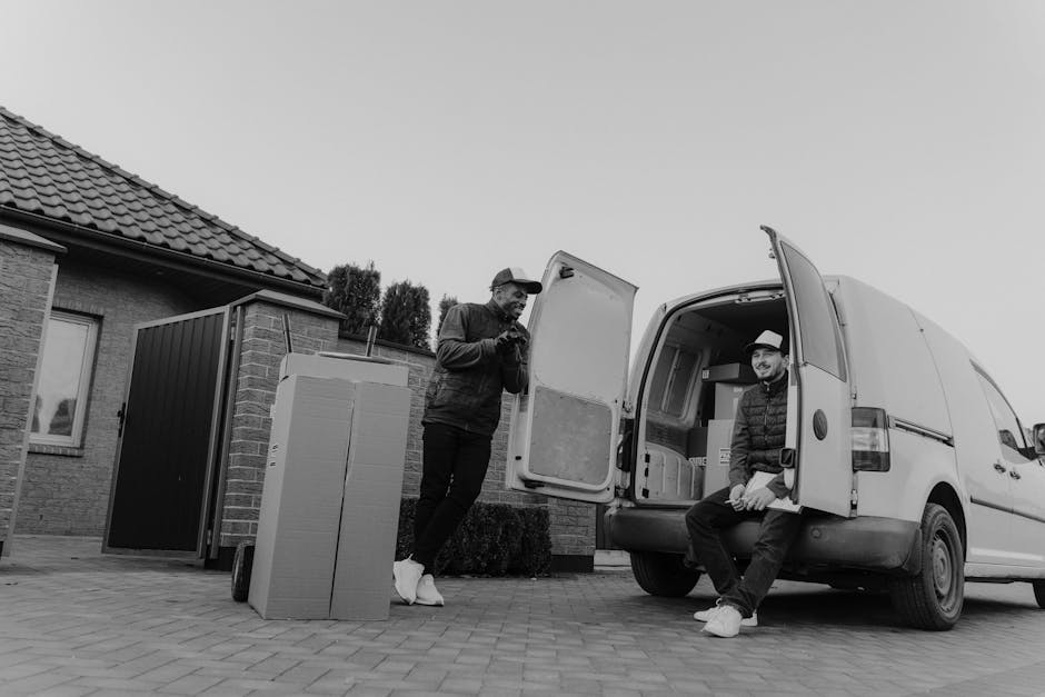Two men involved in a house removal process in a residential driveway. One man, standing outside, is dressed in casual clothing with a jacket and cap, and is holding a small tool or device while looking down. The second man is sitting on the open rear door of a white van, which is parked close to a brick house with a tiled roof. The vehicle's loading area is visible, containing cardboard boxes, foam packaging, and protected furniture wrapped in blankets. Nearby, a hand truck or dolly is positioned with a large piece of furniture or boxed item secured on it. The scene depicts an organized loading process, with the van positioned for furniture transport and packing materials arranged outside. The environment includes a paved driveway, the brick exterior of the house, and some bushes, all under natural daylight. The image emphasizes professionalism and efficiency in home relocation, with Man and Van Highbury providing furniture transport and moving services.