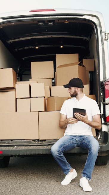 Two men involved in a house removal process in a residential driveway. One man, standing outside, is dressed in casual clothing with a jacket and cap, and is holding a small tool or device while looking down. The second man is sitting on the open rear door of a white van, which is parked close to a brick house with a tiled roof. The vehicle's loading area is visible, containing cardboard boxes, foam packaging, and protected furniture wrapped in blankets. Nearby, a hand truck or dolly is positioned with a large piece of furniture or boxed item secured on it. The scene depicts an organized loading process, with the van positioned for furniture transport and packing materials arranged outside. The environment includes a paved driveway, the brick exterior of the house, and some bushes, all under natural daylight. The image emphasizes professionalism and efficiency in home relocation, with Man and Van Highbury providing furniture transport and moving services.