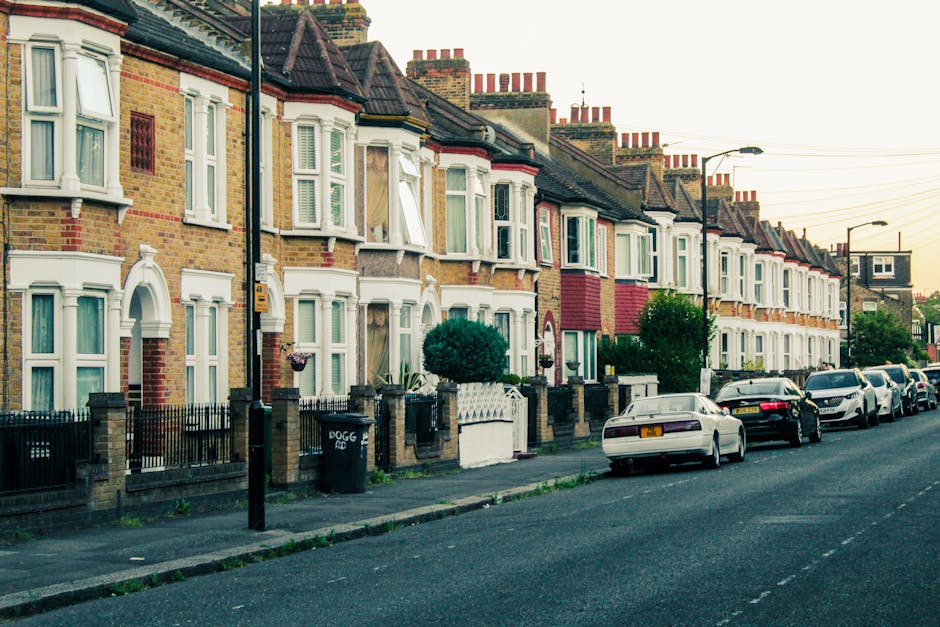 A row of terraced houses with brick facades and white bay windows lining a residential street in Highbury N5. Some windows are open, revealing interior curtains or blinds, while others are closed. The front gardens are enclosed by low brick walls and black metal fences, with small trees and shrubs visible in some gardens. Several cars are parked along the pavement, including a white vehicle closest to the camera. Street lamps and utility poles with overhead wires run parallel to the road. The image captures a quiet, suburban scene during daylight, illustrating the typical housing and street scene connected to house removals, furniture transport, and home relocation services. The environment appears clean and well-maintained, with a focus on the exterior of the properties and the street environment where moving logistics would be carried out.