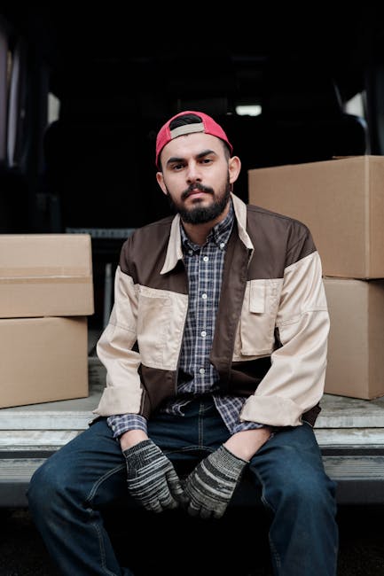 A young man with a beard, wearing a red baseball cap backwards, a checkered shirt, beige and brown work jacket, and gloves, is sitting on the tailgate of a black van during a home relocation. The van's interior is visible behind him, showing stacked cardboard boxes of various sizes ready for packing and transport. The man appears to be taking a break from loading or unloading, with some boxes positioned on the pavement beside him. The scene is set outdoors, possibly on a driveway or street, with natural lighting indicating daytime. This image reflects the process of furniture transport and packing in a professional house removal service, as provided by Man and Van Highbury, supporting accurate and accessible visual depiction of moving logistics.