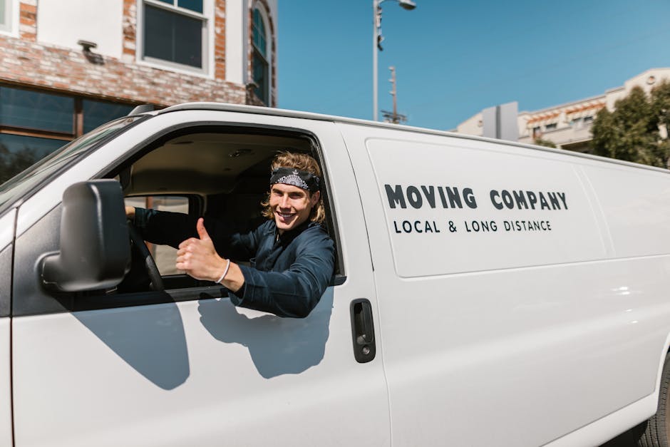 A young male mover with shoulder-length hair, wearing a black bandana and a dark blue work shirt, is sitting inside a white moving van parked on a residential street during daytime. He is smiling and giving a thumbs-up through the open driver's side window. Inside the van, various packing materials such as cardboard boxes, plastic wrapping, and blankets are visible, suggesting a home relocation process. The van bears the text 'MOVING COMPANY LOCAL & LONG DISTANCE' printed in bold black lettering on its side. Behind the vehicle, there are modern multi-storey buildings with brick and glass facades, and a clear blue sky overhead. The scene depicts the loading or unloading phase typical of professional removals by Man and Van Highbury, supporting services related to furniture transport and packing for house moves.