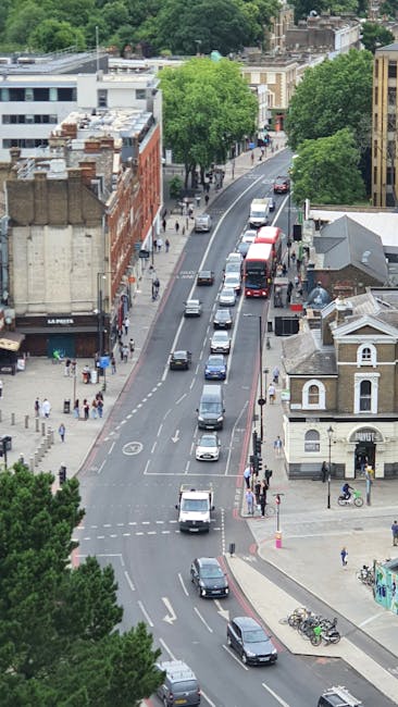 An aerial view of a busy street near Highbury and Islington Station featuring a row of parked cars along the curb and several moving vehicles on the road. On the right side, a small white building with a pitched roof and arched windows is visible, along with a bicycle rack holding multiple bikes. To the left, there are commercial buildings with flat roofs, some with outdoor signage, and pedestrians walking along the sidewalks. Several trees with lush green foliage line the street, providing shade to the pedestrians. The scene captures a typical urban environment during daytime, relevant to house and office removals, illustrating the logistical challenges of furniture transport and packing and moving processes within a city setting. Man and Van Highbury's services could be involved in navigating such environments for efficient home relocation or office removals, ensuring safe loading, packing, and moving of furniture and boxes through busy urban streets.