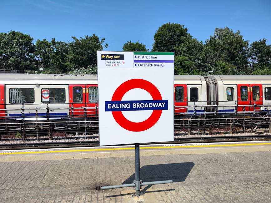 A large white sign with the London Underground roundel symbol and the text 'Ealing Broadway' is positioned on a paved platform at Ealing Broadway station. The sign provides directional information, featuring arrows pointing towards exits, district line, Elizabeth line, national rail, and bus services. Behind the sign, a modern train with a silver body and orange and blue accents is stationary on the tracks, with several closed windows visible along its length. The platform is bordered by a metal railing, and the background includes lush green trees under a clear blue sky, indicating a bright, sunny day. The environment suggests a busy station area where public transportation and train services are operational, with the setting potentially relevant for house removals or moving logistics involving transport connections managed by companies like Man and Van Highbury.