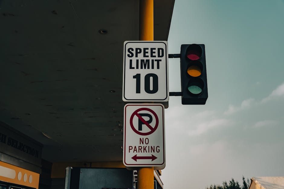 A vertical street sign mounted on a yellow pole under a gray overhang, displaying a speed limit of 10 miles per hour with a traffic light showing red, yellow, and green signals. Below the speed limit