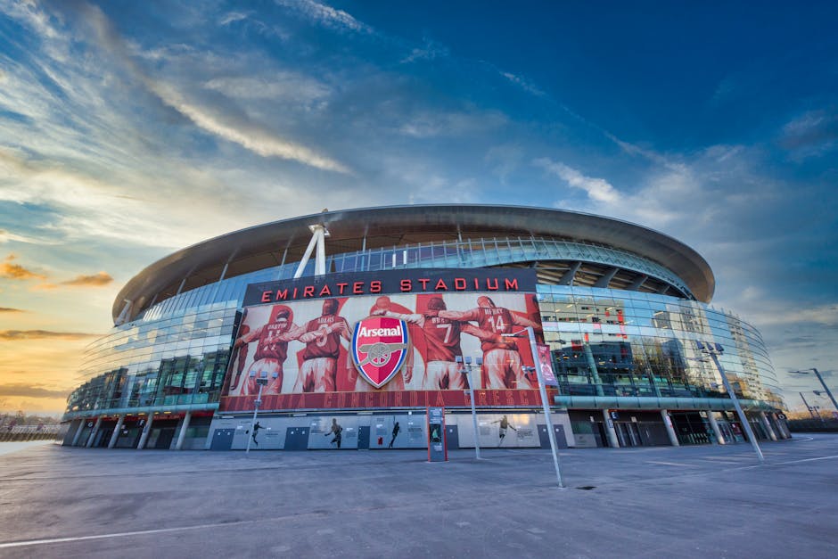 A wide-angle view of Emirates Stadium in Highbury, showing the modern glass facade and the large digital screen displaying Arsenal football players with the club's crest. The stadium is illuminated by natural light during sunset, with partly cloudy skies in the background. In front of the stadium, there is an open paved area with multiple empty parking spaces and a few pedestrians walking nearby. The scene reflects a clean, well-maintained property ready for home relocation or moving services, which Man and Van Highbury offers as part of their removals and furniture transport solutions. The image captures the environment around the stadium, emphasizing spaciousness suitable for loading and unloading furniture, boxes, and packaging materials during a professional removal process.