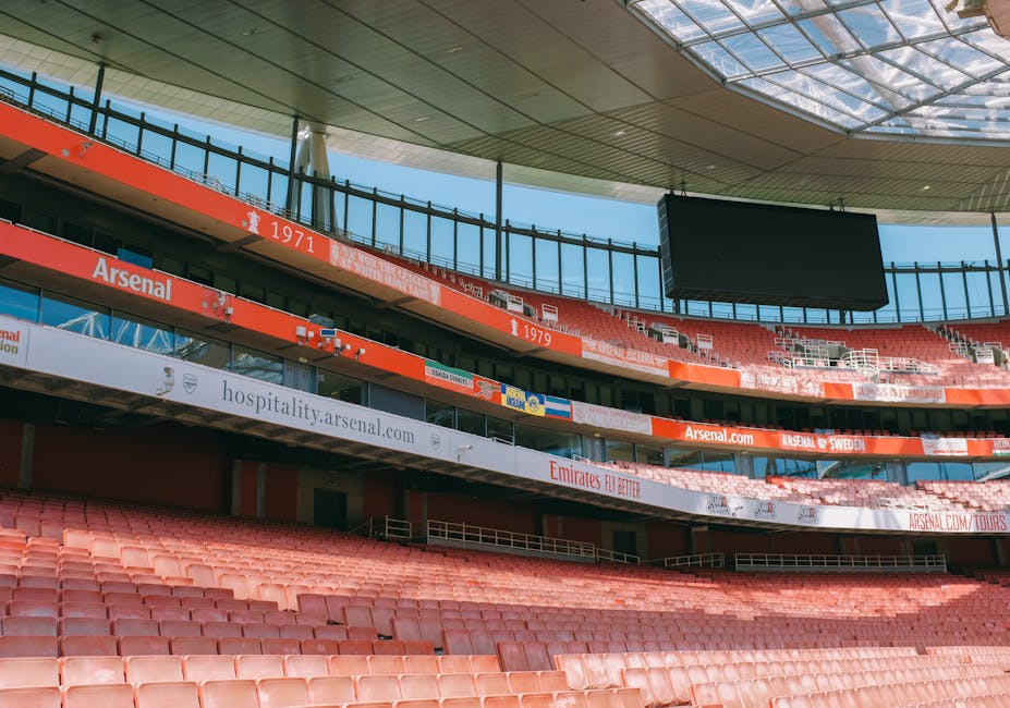 Inside a partially empty outdoor stadium, rows of red plastic seats are visible, with some sections covered by a curved roof structure, and an empty large black digital screen hanging from the ceiling. The stadium has multiple levels of seating separated by white and red advertising banners displaying URLs and sponsorships, including references to Arsenal football club and Emirates. Bright daylight illuminates the scene, casting shadows on the seats and highlighting the architectural features of the upper levels with glass railings. The image captures the interior environment of a sports arena, focusing on the seating area and structural elements, relevant to house removals and furniture transport service providers such as Man and Van Highbury, supporting efficient home relocation logistics.