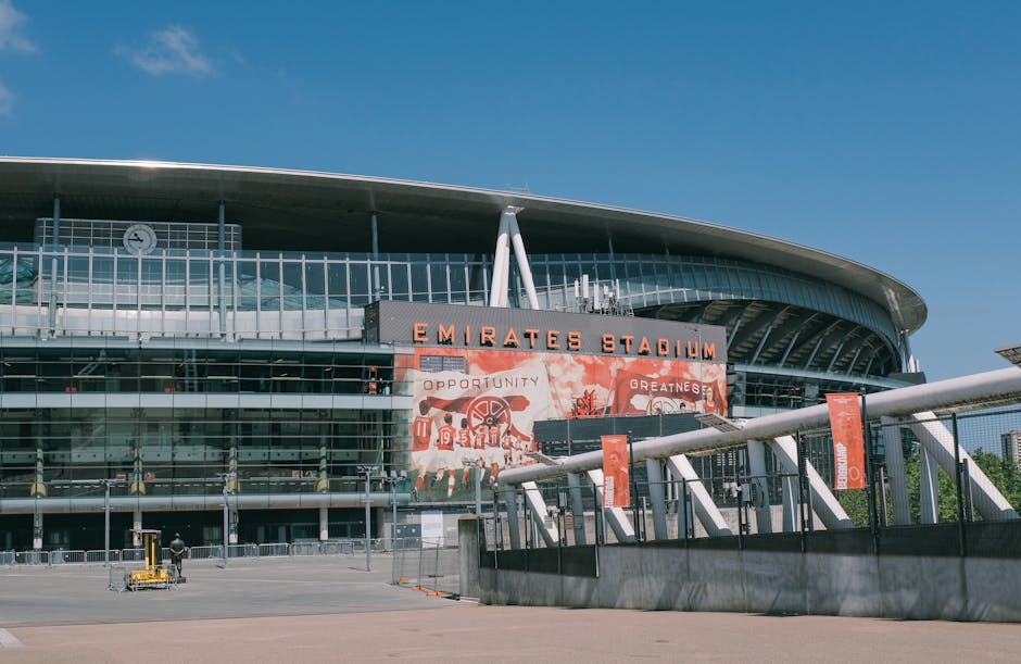A wide-angle view of Emirates Stadium in Highbury, showing the modern glass facade and the large digital screen displaying Arsenal football players with the club's crest. The stadium is illuminated by natural light during sunset, with partly cloudy skies in the background. In front of the stadium, there is an open paved area with multiple empty parking spaces and a few pedestrians walking nearby. The scene reflects a clean, well-maintained property ready for home relocation or moving services, which Man and Van Highbury offers as part of their removals and furniture transport solutions. The image captures the environment around the stadium, emphasizing spaciousness suitable for loading and unloading furniture, boxes, and packaging materials during a professional removal process.