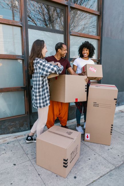 Three individuals are standing outside a building with large windowpanes framed by wooden mullions, engaged in a home relocation process with packing boxes. The first person, a woman with long brown hair wearing a blue and white checkered shirt and light-colored trousers, is holding a medium-sized cardboard box. The second person, a man with short dark hair dressed in an orange pair of trousers and a dark red t-shirt, is also holding a box, preparing to load or unload. The third person, a woman with curly black hair wearing a white t-shirt and blue jeans, is smiling and holding a smaller cardboard box with red and white labels indicating contents. Several boxes of different sizes, some with printed labels, are positioned on the ground and held by the individuals, suggesting the process of packing or unpacking for a house move. The scene is set on a paved sidewalk during daytime, with natural light illuminating the area, capturing elements typical of professional removals and furniture transport, aligned with services offered by Man and Van Highbury.